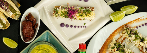 A top-down view of assorted Mexican dishes, including tacos, enchiladas, guacamole, limes, and a bottle of hot sauce on white and green plates, garnished with herbs and purple sauce dots.