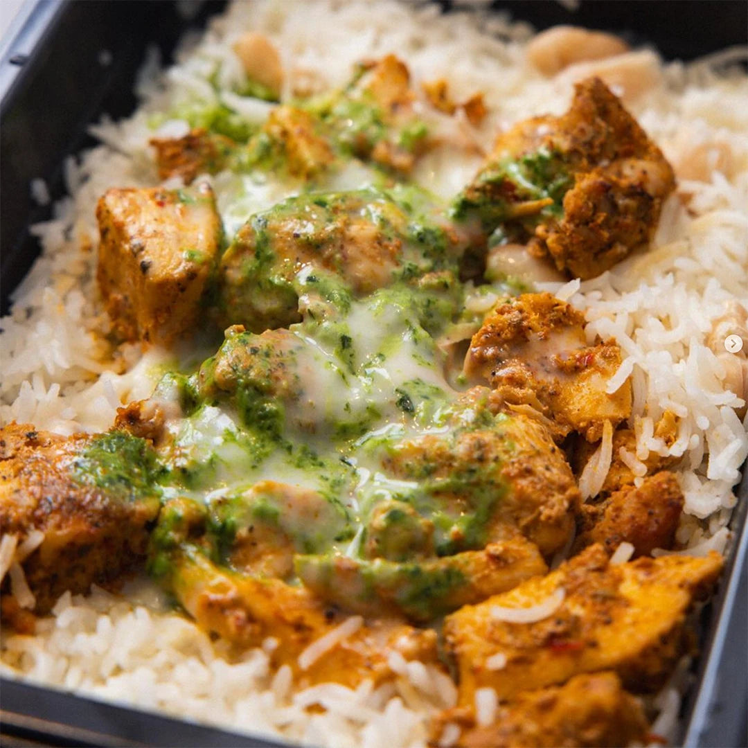 A close-up of spiced grilled chicken pieces on a bed of white rice, topped with creamy green and white sauces, served in a black container.