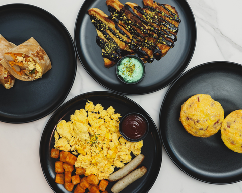 Flat lay of a black plate breakfast spread with a halved burrito, chocolate-drizzled French toast with pistachios and green dip, scrambled eggs, sausage, hash browns with ketchup, and two bacon egg muffins on a white marble surface.