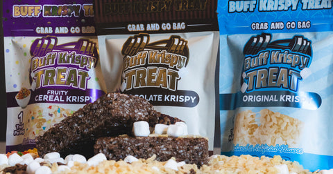 Three Buff Krispy Treat flavors—Fruity Cereal Krispy, Chocolate Krispy, and Original Krispy—are displayed in colorful "Grab and Go" bags behind stacked chocolate treats topped with mini marshmallows, surrounded by rice crisps.