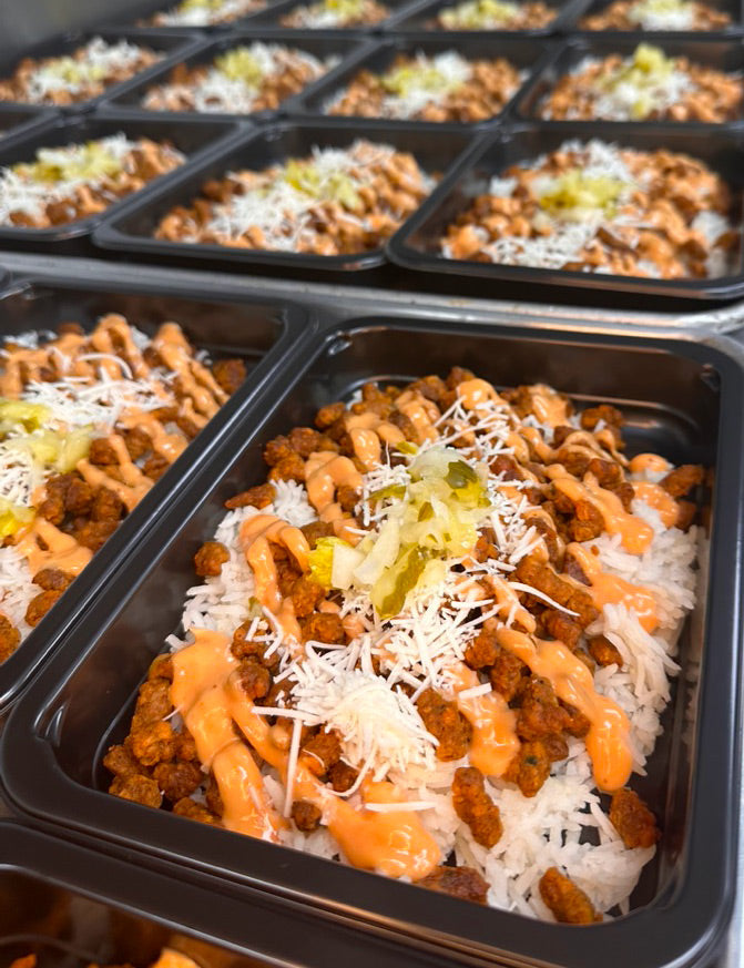 Multiple black meal prep containers filled with white rice, seasoned ground meat, shredded cheese, a drizzle of orange sauce, and a garnish of pickles are arranged in rows.