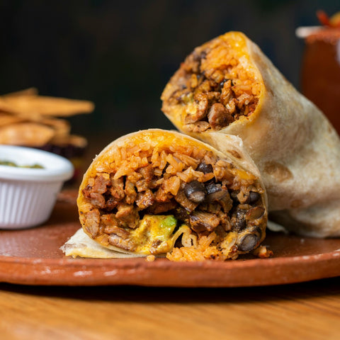 A close-up of a burrito cut in half, showing its filling of rice, beans, meat, and vegetables, on a brown plate with fries and a small cup of sauce in the background.