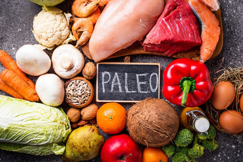 A variety of fresh foods, including vegetables, fruits, shrimp, eggs, seeds, and nuts, surround a small chalkboard with PALEO written on it, representing foods allowed on the Paleo diet.