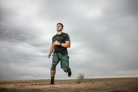 A man in athletic clothing runs on sandy terrain under a cloudy sky, with dust rising behind him as he moves forward.