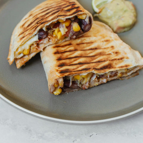 Two grilled quesadilla halves filled with black beans, corn, and cheese on a gray plate, served with a dollop of guacamole and a lime wedge in the background.