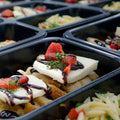 Close-up of meal trays containing breaded chicken topped with melted cheese, a drizzle of balsamic glaze, diced red peppers, and herbs, with a side of pasta salad in a black plastic container.