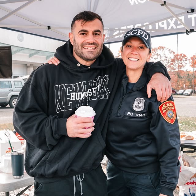 A man in a black hoodie and a woman in an SCPD police uniform smile with their arms around each other under a tent, with food and drinks on the table behind them.