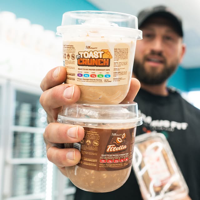 A man holds up two containers of overnight oats, labeled Toast Crunch and Fitella, displaying their packaging toward the camera in a brightly lit store.