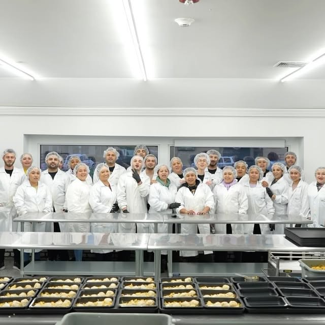A group of people in white lab coats and hairnets pose for a group photo in a bright, clean kitchen or food production area with trays of pastries or dough on racks in the foreground.