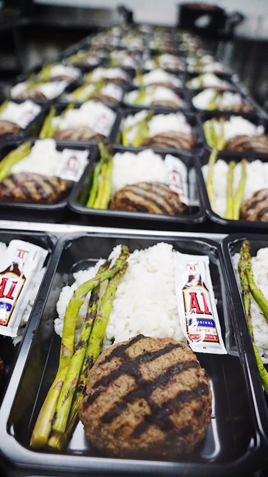 Rows of black meal prep containers filled with grilled asparagus, white rice, a hamburger patty, and a packet of A1 sauce are lined up on a table, ready for distribution.