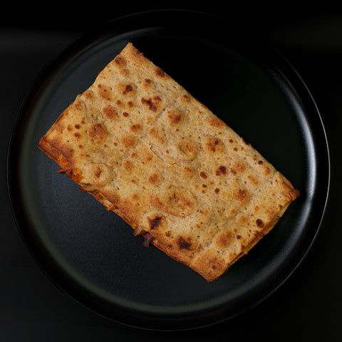 The Strongsville Chicken Ranch Fold is a golden brown rectangular flatbread with dark toasted spots, served on a round black plate against a black background—ideal for high protein meals or chicken ranch folds.