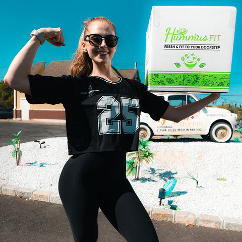 A smiling woman in sunglasses and a Women’s Cropped Mesh Jersey – Black White Trim flexes her arm and holds a Hummus Fit delivery box outdoors, with a branded truck and bright blue sky in the background.
