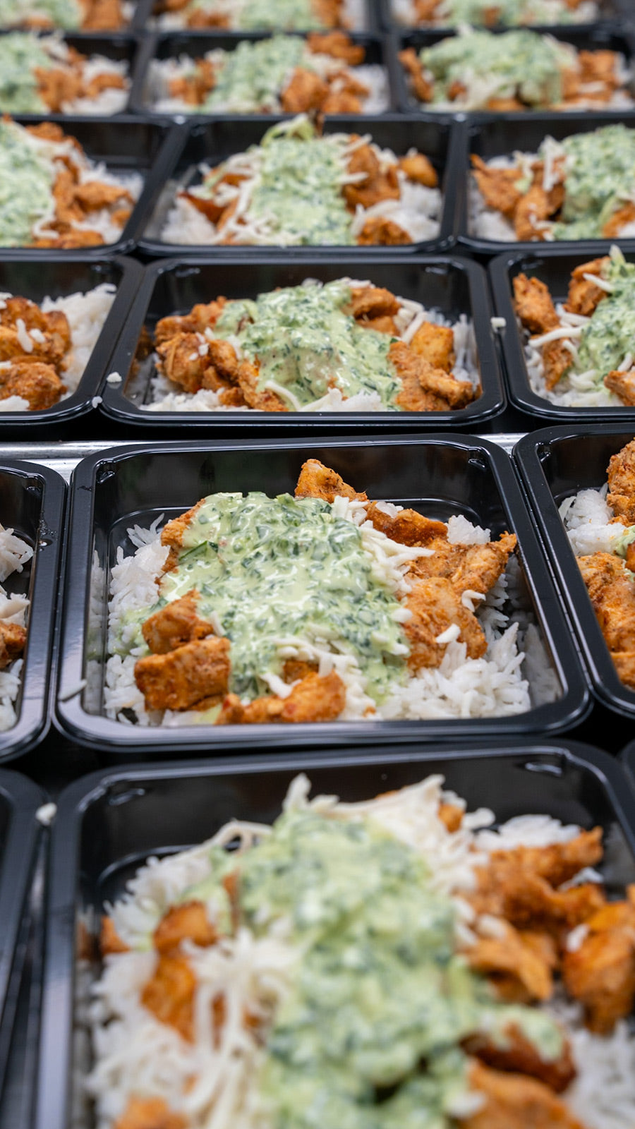 Several black meal prep containers filled with white rice, spiced chicken pieces, and topped with a creamy green herb sauce are arranged in neat rows.