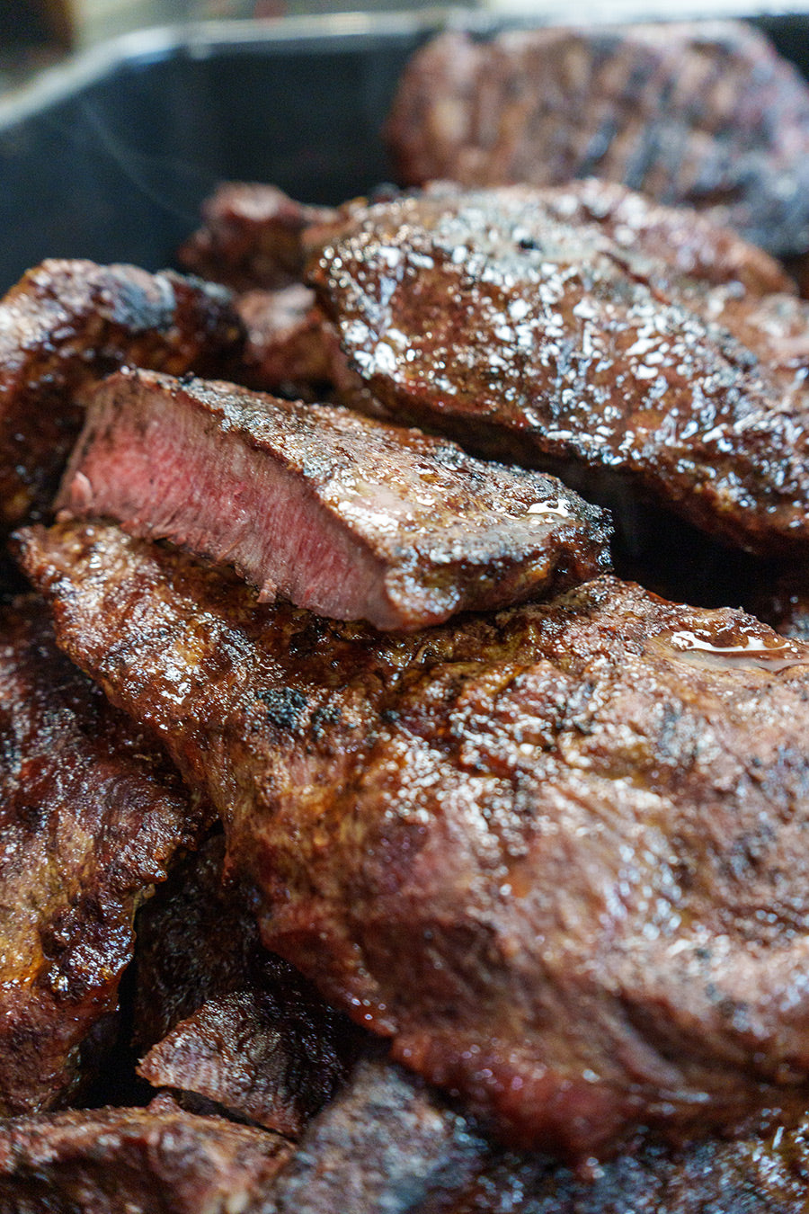 Close-up of several grilled steaks stacked together, with one piece cut to reveal a juicy, medium-rare pink center. The meat is glistening and has a charred, seasoned exterior.