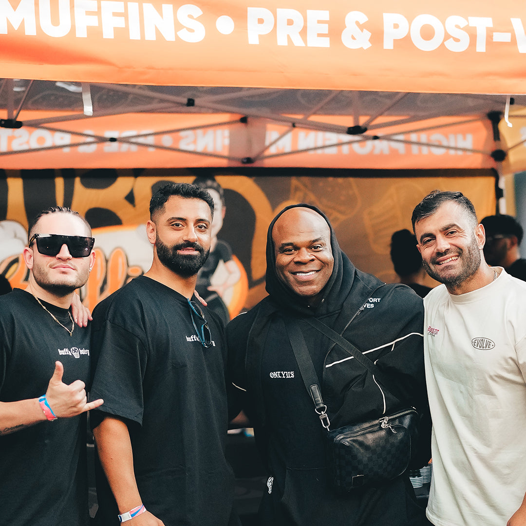 Four men smiling and posing together under an orange event tent with white text. One man wears sunglasses, another wears a hoodie and crossbody bag, and the others are dressed casually. The background shows more people and event signage.