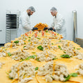 Two people in white lab coats and hairnets assemble sandwiches on a long table covered with tortillas, greens, and shredded meat in a commercial kitchen. A tall stack of sandwich buns is visible between them.