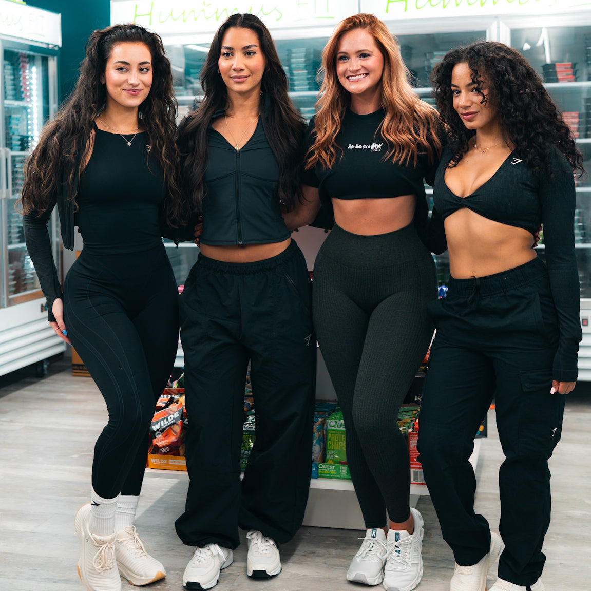 Four women in athletic wear stand together and smile inside a store. Shelves of products and a sign reading “Humans Fit” are visible in the background. They appear happy and relaxed, posing for the photo.