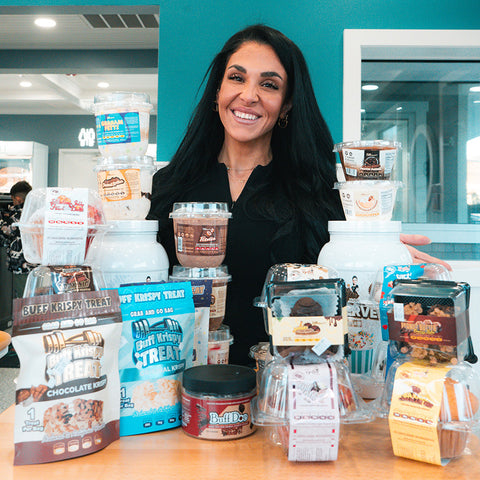 Woman standing behind a display of protein powder and snack products in a store setting.