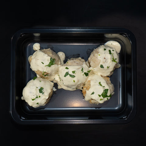 Four Keto Ricotta Meatballs made with ground chicken, topped with creamy white sauce and herbs, are served in a black rectangular tray on a dark background for a tasty low-carb meal.