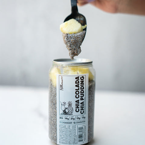 A hand holds a spoonful of Chia Colada Chia Pudding topped with pineapple over a labeled jar, showing layers of chia pudding and fruit—ideal for healthy meal prep.