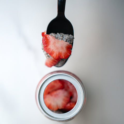 A black spoon holds a scoop of Jammin Chia Pudding (Subscription) topped with sliced strawberry above an open jar filled with more strawberries and chia pudding, creating a delicious, healthy snack on a white background.