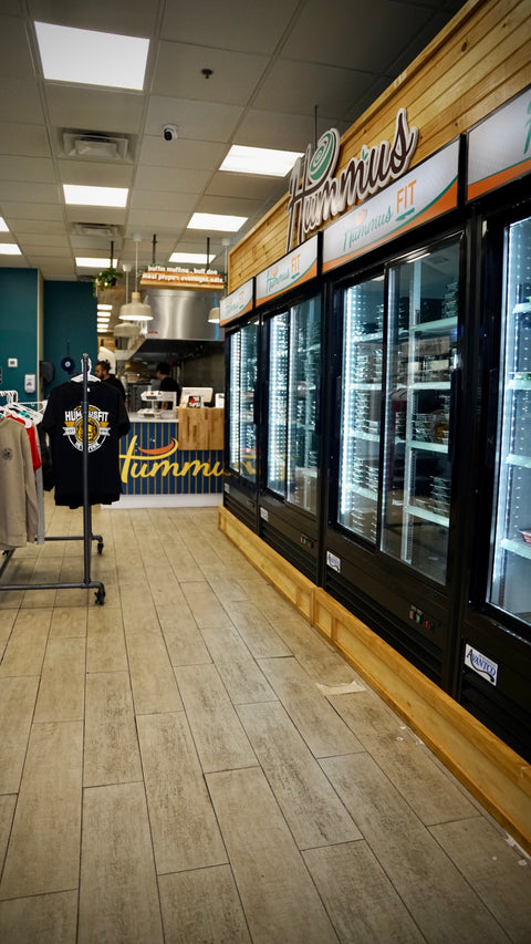 A grocery store aisle with glass-door refrigerators on the right, labeled Hummus FIT. Clothing, including a black t-shirt, is displayed on racks to the left. The store has a light wood floor and bright lighting.