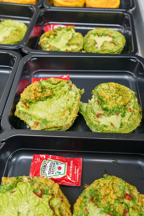 Several black meal trays each contain two green egg muffins with bits of red and green vegetables, accompanied by a packet of Heinz tomato ketchup.