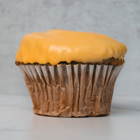 A Buffin Muffin - Buffalicious Baddie, in a crinkled paper liner and crowned with smooth yellow frosting, rests on a light surface against a pale, textured background.
