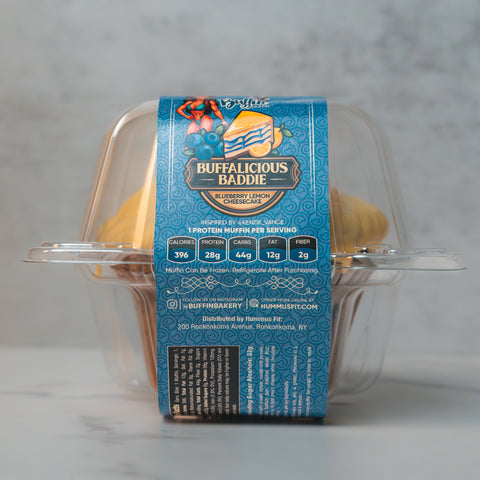 A packaged muffin with a blue patterned label reading “Buffalicious Baddie, Blueberry Lemon Cheesecake.” The label lists calories, protein, carbs, fat, and fiber per serving. The clear plastic container shows the muffin inside.