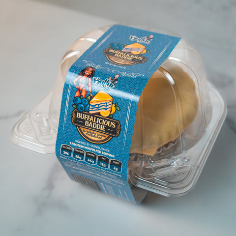 A clear plastic container displays the Buffin Muffin - Buffalicious Baddie with nutrition info and a blue label featuring a cartoon woman, blueberry graphics, and promotes it as a gym-friendly snack.