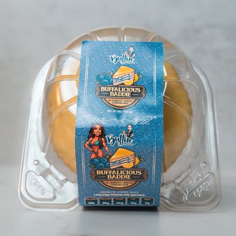 A clear plastic container holds a round yellow cheesecake. The blue label reads Buffin Muffin - Buffalicious Baddie and features illustrated blueberries with a cartoon woman in red—ideal as a gym-friendly snack.
