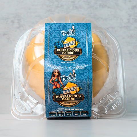 A clear plastic container holds a yellow muffin. The blue label reads Buffalicious Baddie Blueberry Blondie and features images of blueberries and a cartoon woman with red hair. Nutritional info is listed at the bottom.