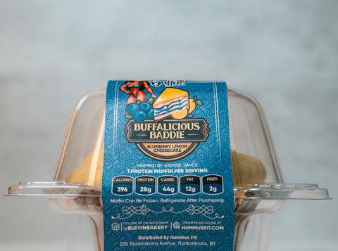 A clear plastic container holds a muffin with a blue label reading Buffalicious Baddie Blueberry Lemon Cheesecake. Nutritional info, website, and distributor details are visible on the label.
