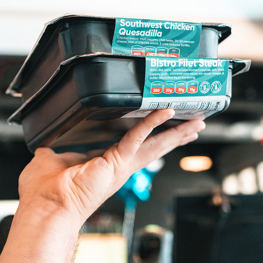 A hand holds up two stacked black meal containers with teal labels reading Southwest Chicken Quesadilla and Bistro Filet Steak in a bright indoor setting.