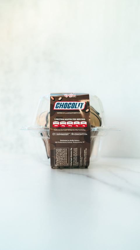 A clear plastic container holds a chocolate muffin with a brown label reading CHOCOLIT EXCLUSIVE and listing nutritional facts. The package rests on a white marble surface with a bright background.