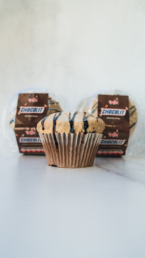 A chocolate cupcake with light icing and chocolate drizzle sits on a white surface, with two packaged CHOCOLIT EXCLUSIVE cupcakes in the background.