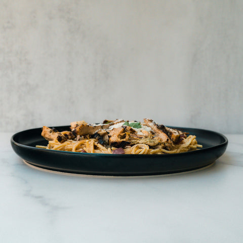 A black plate with Gluten Free Carbonara Chicken Subscription—high-protein pasta topped with grilled chicken, grated cheese, and herbs—set on marble against a neutral backdrop, for perfect Italian comfort food.