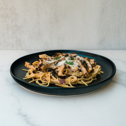 A black plate with Gluten Free Carbonara Chicken Subscription—high-protein pasta, grilled chicken strips, veggies, and grated cheese—rests on a white marble surface against a light backdrop.