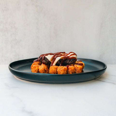 The Everything But the Bagel Burger features crispy tater tots topped with sausage, melted cheese, and BBQ sauce, artfully arranged on a dark plate over light marble with a neutral backdrop.