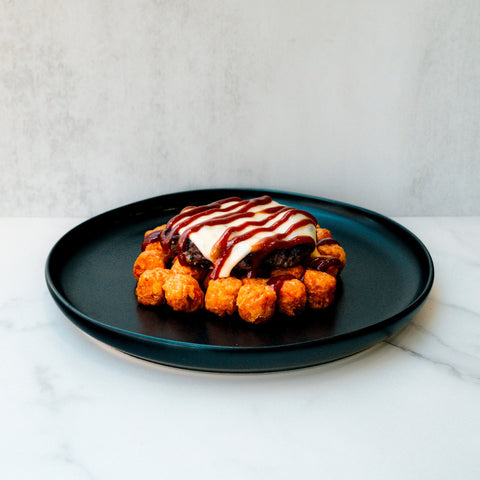 The Everything But the Bagel Burger, topped with melted cheese, sautéed onions, and BBQ sauce, sits on a black plate surrounded by golden tater tots. The plate rests on white marble against a light background.
