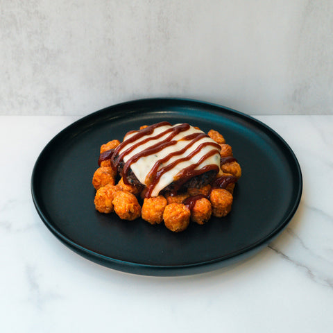 The Everything But the Bagel Burger sits on a black plate, topped with melted cheese and barbecue sauce, surrounded by crispy tater tots, all presented neatly on a white marble surface.