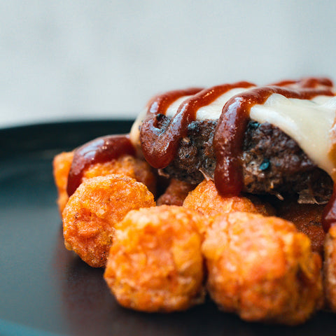 A close-up of the Everything But the Bagel Burger: crispy tater tots topped with a high-protein patty, melted cheese, and barbecue sauce, served on a dark plate.