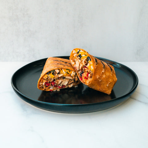 The Hey Arnold Burrito—grilled and cut in half, packed with chicken, cheese, rice, beans, and veggies—is served on a black plate against a light background. It’s the ideal high-protein chicken burrito for your post-workout meal.