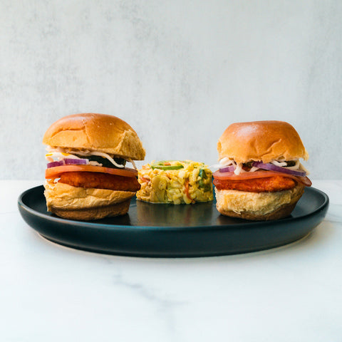 Two Hot Honey Chicken Sliders Subscription, each with crisp lettuce, tomato, onion, and a breaded patty for a swicy kick and high-protein boost, are served on a black plate with a mixed vegetable salad in the center.