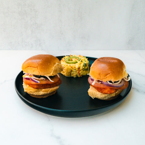 Two Hot Honey Chicken Sliders with sliced red onions, tomatoes, and patties are served on a black plate, accompanied by a round portion of mixed vegetable salad against a plain, light background.