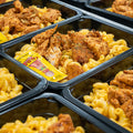 Multiple black meal prep containers filled with macaroni and cheese, fried chicken pieces, and a packet of hot sauce, arranged in rows on a table.