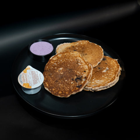 Three pancakes on a black plate with a small cup of blueberry sauce and a container of whipped butter, set against a dark background.
