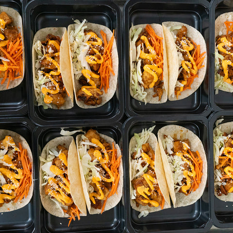 Several meal prep containers each hold two soft tacos filled with shredded cabbage, sliced carrots, seasoned meat, orange sauce, and a sprinkle of sesame seeds, arranged neatly in rows.