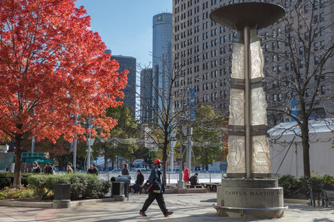 People walk and sit near the illuminated Campus Martius monument in downtown Detroit, surrounded by tall buildings and vibrant autumn trees with red leaves.