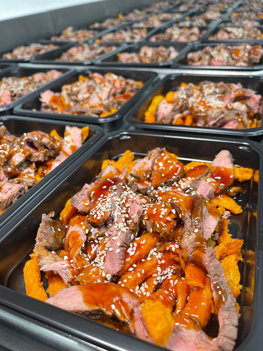 Rows of black meal prep containers filled with sliced steak, sweet potato fries, drizzled sauce, and sprinkled sesame seeds are lined up on a countertop.